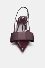 Zapato destalonado de mujer, color borgoña (vino), con acabado acharolado y punta fina. Presenta un lazo de tela en la parte delantera y una tira fina en el talón. El tacón es ancho y de altura media.
