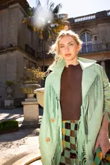Chaqueta de cuero ovino color verde menta, con cuello sastre, cierre frontal de botones y flecos decorativos en las solapas, mangas y puños. Presenta un diseño holgado y bolsillos laterales.
