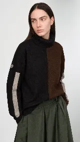 Polera de cuello alto confeccionada en lana lambswool, con diseño bicolor en negro y marrón. Presenta mangas largas con franjas verticales en gris y un parche circular bordado en la manga derecha.