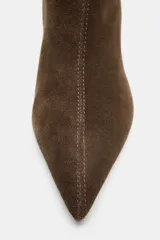 Botín de caña media en piel de ante color marrón, con diseño de punta fina y tacón alto tipo aguja. Presenta costuras verticales en el cuerpo y una estructura tipo pull-on para facilitar el calce.