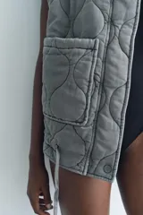 Chaleco gris acolchado con cuello alto, cierre frontal con cremallera metálica, bolsillos de plastrón en delantero y bajo ajustable con cordones.