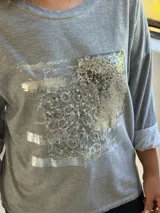 Remera gris de manga larga con cuello redondo y diseño holgado. Presenta un aplique frontal rectangular con detalles de lentejuelas plateadas y estampado tipográfico.