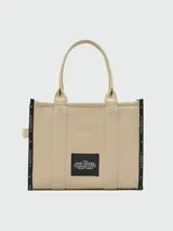 Bolso tipo tote de Marc Jacobs, modelo The Jacquard Large Tote Bag, confeccionado en tejido jacquard color tostado. Presenta el nombre de la marca y el modelo bordados en el frente en color blanco, doble asa superior y detalles de cintas laterales con inscripciones.