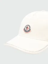 Gorra de baseball color blanco crudo, con visera curva y costuras decorativas en zigzag a contratono en el borde. Presenta un parche circular bordado con el logo de la marca en el frente.