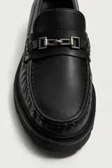 Mocasín de vestir negro con parte superior lisa, punta redonda, suela track y detalle de aplique metálico en el empeine.