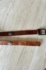 Cinturón de cuero vacuno color cognac con hebilla metálica rectangular, decorado con tachas pequeñas y apliques metálicos circulares grabados.