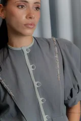 Chaqueta gris de silueta femenina con mangas abullonadas, cuello redondo y cierre frontal con botones forrados. Presenta detalles de bordado artesanal a mano en los hombros y solapas.