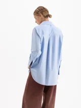 Camisa de corte holgado confeccionada en poplín de algodón. Presenta cuello clásico con interior en tela a rayas a contratono, cierre frontal con botones al tono, bolsillo plaque en el pecho con detalle de remache metálico en forma de corazón y mangas largas con puños estructurados.