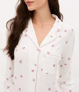 Pijama de dos piezas estilo americano, compuesto por camisa de manga larga con cuello en V, botones frontales y bolsillo en el pecho, y pantalón largo con cintura elástica. Presenta un estampado floral delicado sobre fondo blanco y detalles de encaje rosado en los puños y el ruedo del pantalón.