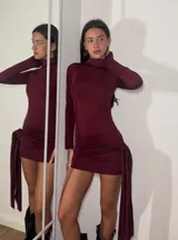 Vestido corto de cuello alto y manga larga, confeccionado en tejido elástico color bordó. Presenta un diseño ajustado al cuerpo con un detalle de nudo lateral y caída de tela asimétrica.