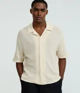 Camisa masculina de manga corta, color crema, con cuello bowling y calce boxy. El tejido presenta un efecto punto texturizado.