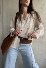 Camisa de manga larga color beige con detalles de encaje y calados verticales en el frente y las mangas, cuello en V y cierre frontal con botones.