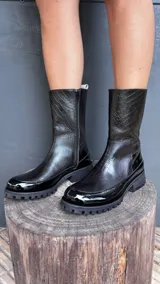 Bota de caña 3/4 color negro, con textura de cocodrilo en la parte superior y acabado charol en la puntera y el talón. Tiene cierre lateral y suela track.
