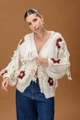 Saco de punto grueso color crudo, con diseño de calce relajado y mangas amplias. Presenta apliques tejidos con motivos florales en color terracota y detalles de tiras anudadas decorativas en toda la prenda.