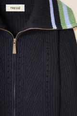 Cárdigan de punto azul marino con cierre frontal de cremallera. Presenta cuello tipo camionero y puños con detalles de rayas en tonos verde y celeste. El tejido tiene una textura acanalada con detalles calados decorativos.