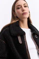 Campera tipo teddy de textura suave y acabado de peluche en color beige. Presenta cuello amplio con detalle de hebilla, cierre frontal y recortes en contraste de color marrón que marcan la estructura de la prenda.