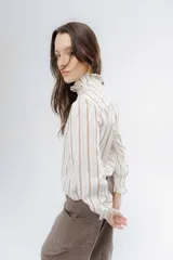 Camisa de corte clásico con estampado de rayas verticales en tonos neutros. Presenta cuello alto con volados delicados y mangas largas con puños elásticos terminados en volados.