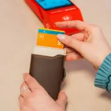 Billetera tarjetero automática con estructura de aluminio y revestimiento de cuero bicolor en tonos verde oliva y beige. Cuenta con un cierre de broche a presión y el logo de la marca grabado en la parte inferior.