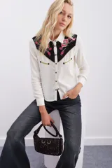 Camisa de estilo western confeccionada en viscosa color crudo. Presenta un canesú delantero y trasero en contraste color negro con bordados florales en tonos fucsia y verde, realzado con vivos en lurex dorado. Cuenta con cierre frontal mediante broches metálicos, mangas largas con puños abotonados y detalles de bolsillos decorativos en el pecho.