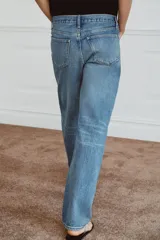 Jeans de tiro medio con corte recto y largo completo. Presentan un diseño de cinco bolsillos, cierre frontal con cremallera y botón, y un acabado con efecto lavado. Incluyen detalles de roturas y parches decorativos en la parte delantera.