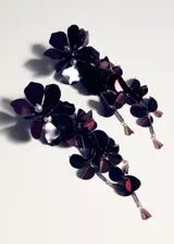 Aros colgantes largos de diseño floral en color violeta oscuro. Presentan múltiples capas de pétalos metálicos y acrílicos, adornados con pequeñas perlas y cristales colgantes en los extremos.