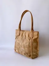 Cartera tipo tote de cuero color camel, con diseño artesanal que presenta costuras decorativas visibles en los laterales y el frente. Cuenta con doble asa de hombro y cierre superior.