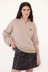 Sweater de tejido acanalado en color beige, con cuello alto y cierre frontal parcial. Presenta un diseño holgado con mangas largas y un pequeño parche con logo en el pecho.