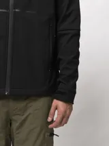 Campera softshell negra con capucha ajustable, cierre frontal completo y bolsillos laterales con cierre. Presenta puños con ajuste de velcro y logo de la marca estampado en el pecho.