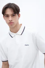 Remera polo blanca con cuello y puños acanalados con rayas azules y rojas. Presenta una media cartera de botones y un bordado en el pecho con la palabra "Uniform" en azul.