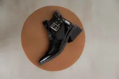 Botineta de cuero charol negro con acabado brillante, diseño de punta fina y taco cuadrado. Presenta un panel elástico lateral y una correa decorativa con hebilla metálica plateada.