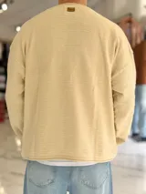 Sweater de punto liviano color beige, con textura marcada, cuello redondo y mangas largas de corte relajado, con pequeño estampado tipográfico negro en el lateral inferior.