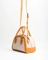 Cartera tipo baúl color tostado con cuerpo de lona y detalles en cuero sintético, asas de mano y correa larga regulable extraíble.
