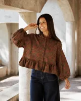 Camisa de lino con estampado floral en tonos tierra y rojizos. Presenta cuello con volado, mangas largas amplias con puños elásticos y un diseño con volado en la cintura que aporta volumen y movimiento.