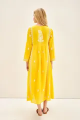 Vestido largo hasta los tobillos de color amarillo, con bordados florales blancos a contratono. Presenta mangas tres cuartos, escote redondo con cartera de botones frontales y un diseño holgado.