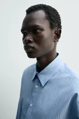 Camisa de algodón celeste con rayas verticales finas en blanco, de corte relajado (relaxed fit), cuello solapa y manga larga con puño abotonado.