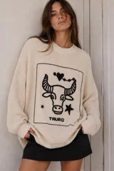 Sweater de punto oversize en color crudo, de manga larga y cuello redondo, con un gran bordado frontal en negro que representa al signo Tauro dentro de un recuadro.