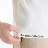 Remera de manga corta New Balance Varsity para hombre, de color blanco marfil con calce relajado y cuello redondo. Presenta un estampado gráfico central con diseño ilustrado y texto bordado en el borde inferior.