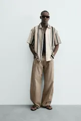 Camisa de manga corta con calce relaxed fit, confeccionada en tejido estructurado con algodón. Presenta un diseño de rayas verticales en tonos beige, negro y blanco, cuello solapa, cierre frontal con botones y aberturas laterales en el bajo.