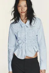 Camisa de popelín de algodón con cuello solapa, manga larga y cierre frontal con botones. Presenta un diseño entallado con detalle de frunces elásticos en el delantero y bajo asimétrico.