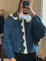 Campera de denim azul con detalles de encaje crudo en el cuello y la solapa frontal, mangas largas con puños elásticos y cierre frontal con botones metálicos dorados.