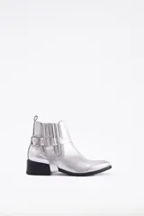 Botín de cuero vacuno en color plateado metalizado, con diseño de caña baja, elásticos laterales y detalle de estribo desmontable con hebilla metálica. Cuenta con un taco cuadrado bajo y suela negra.