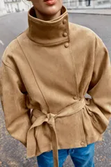 Chaqueta de gamuza sintética color marrón, con cuello subido, cierre frontal cruzado con botones y mangas largas anchas. Cuenta con bolsillos delanteros y cinturón ajustable del mismo tejido.