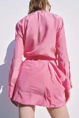 Blusa fluida de color rosado con cuello redondo ajustable mediante lazadas del mismo tejido, manga larga con puño y bajo asimétrico con aberturas laterales.