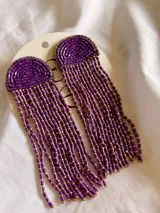 Aros colgantes de diseño artesanal, compuestos por una base semicircular bordada con mostacillas violetas y flecos largos formados por hileras de mostacillas violetas y doradas.