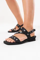Sandalia baja de cuero negro con tiras cruzadas en la capellada decoradas con tachas metálicas plateadas. Cuenta con cierre de hebilla en el talón, suela plana con pequeño tacón y punta cuadrada.