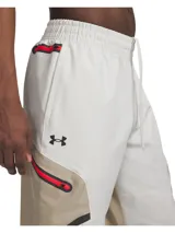 Pantalón cargo técnico de Under Armour, confeccionado en tejido elástico con tecnología UA Storm repelente al agua. Presenta un diseño en tonos beige y blanco con paneles en contraste, bolsillos laterales con cremallera, cintura elástica con cordón interno y bajos ajustables con cordón elástico.