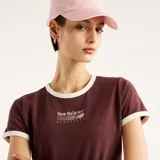 Remera New Balance de mujer, modelo Trackside Ringer, en color borravino con cuello redondo y ribetes en contraste color blanco. Presenta mangas cortas y un gráfico bordado en el centro del pecho.