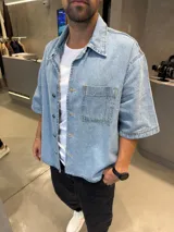 Camisa de jean de corte boxy, color azul claro, con mangas cortas, cuello clásico y cierre frontal con botones metálicos. Presenta un bolsillo frontal en el pecho.