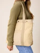 Bolso tipo tote de diseño acolchado con patrón de rombos, confeccionado en poliamida negra. Presenta un bolsillo frontal central de pana con el logo de la marca bordado en dorado, detalles de ribetes en pana negra y botones metálicos dorados en la parte superior. Incluye correas de hombro resistentes y forro interior de algodón a cuadros.