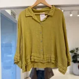 Camisa de lino color verde oliva, de corte oversize y corto, con cuello camisero, cierre frontal con botones y detalles de volados superpuestos en el ruedo y los puños de las mangas largas.